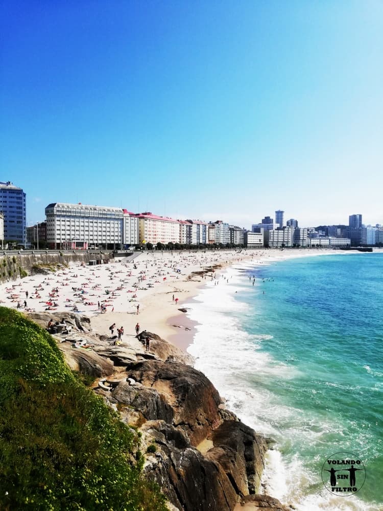 Imagen de la plaza del Orzan desde lo alto del Paseo Marítimo, se ve el mar azul y personas en la playa en un día despejado