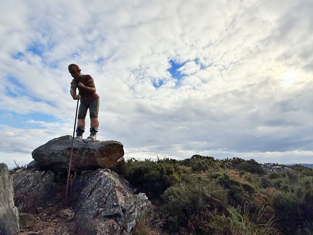 Se ve una la escultura del Mouro con todo el cielo con nubes en la parte de atrás. Es una figura de persona con un palo sobre una roca alta 