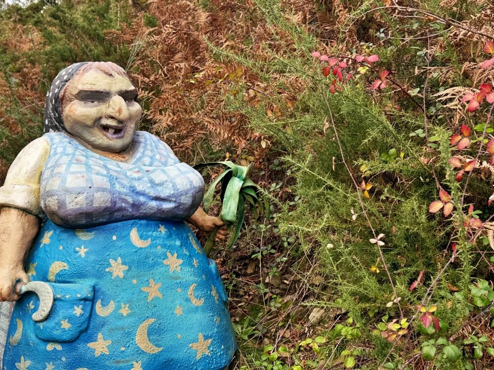 Se ve en primer plano la estatua de la Meiga, que lleva una falda azul con estrellas y lunas y una pañoleta 